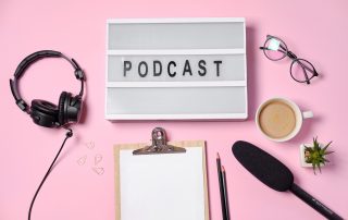 Auf einem rosa Untergrund liegt Podcastequipment, sowie eine Brille, ein Notizblock und ein Kaffee.